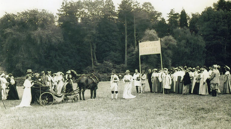 A black and white Edwardian photograph of people gathered on a field, holding a banner, in contemporary dress. A horse and cart is also there.